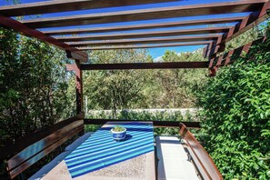 Une pergola en bois surmontée d’un tapis à rayures bleues et blanches, avec un banc et un pot de fleurs.