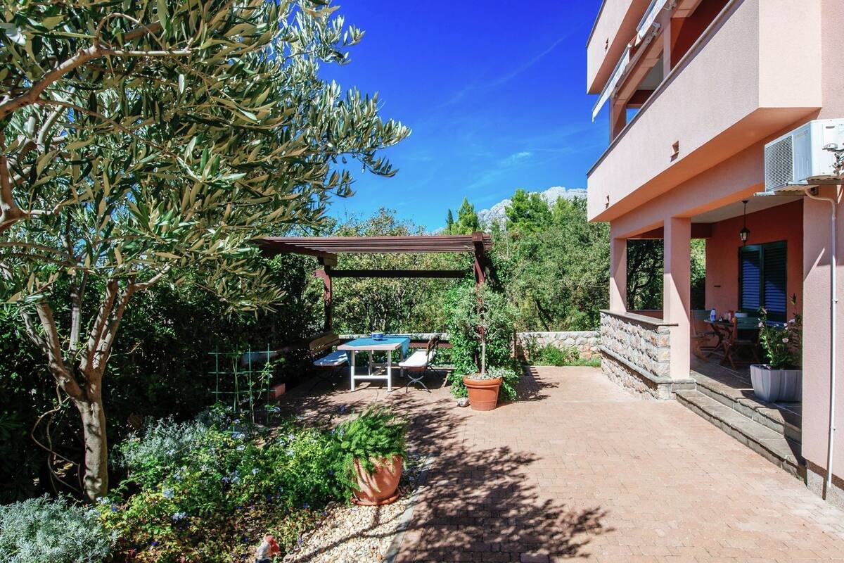 A covered outdoor seating area with tables and chairs, surrounded by potted plants and a pergola.