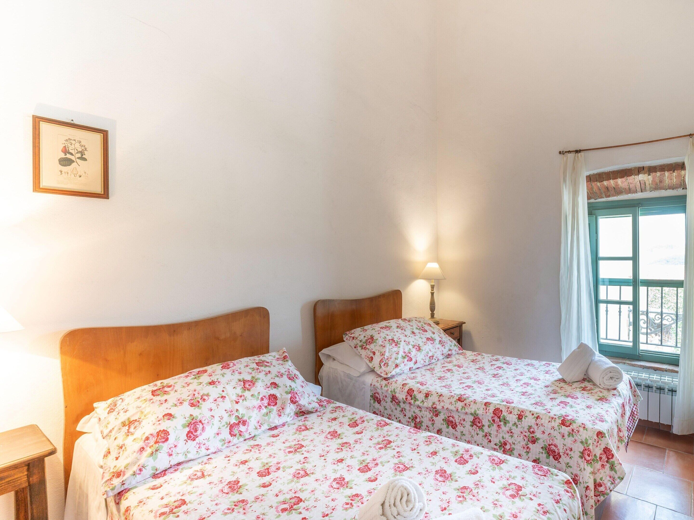 Una habitación con dos camas, una mesita de noche con lámpara, una ilustración botánica enmarcada en la pared y una ventana con cortinas.