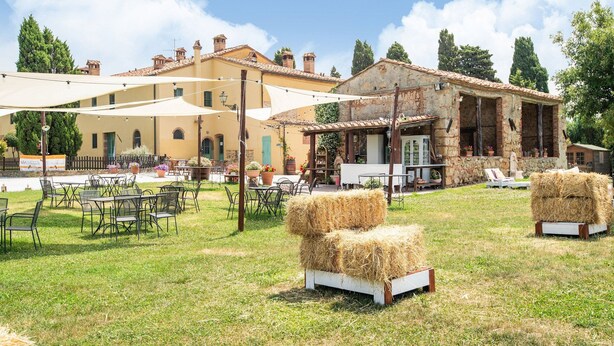 An outdoor seating area with tables and chairs, hay bales, and a building with stone walls and a tiled roof. - Appealing Apartment in Collesalvetti With Garden (Collesalvetti)