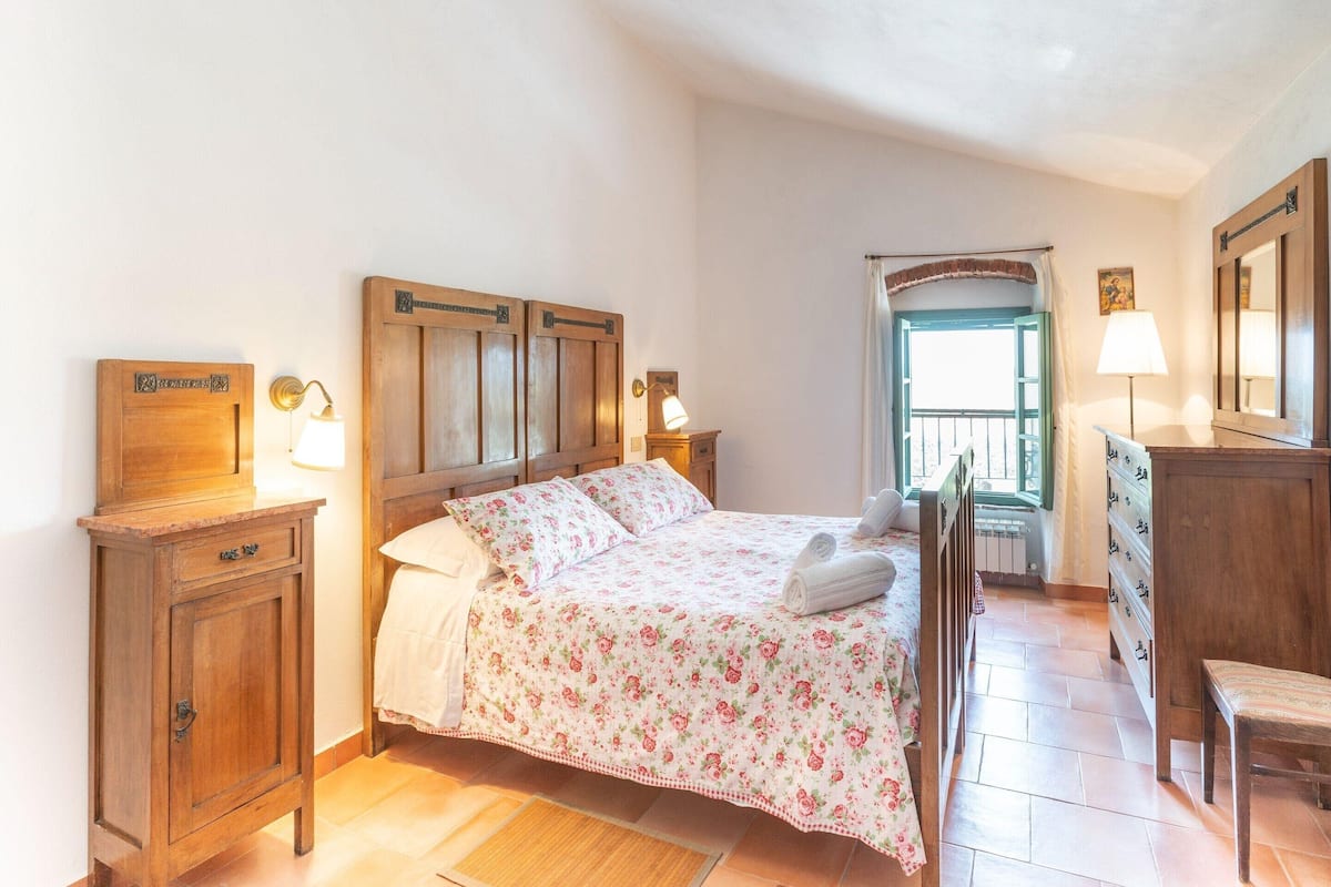 A bedroom with a wooden bed, two bedside tables, a nightstand, a dresser, a chair, and a window with curtains.