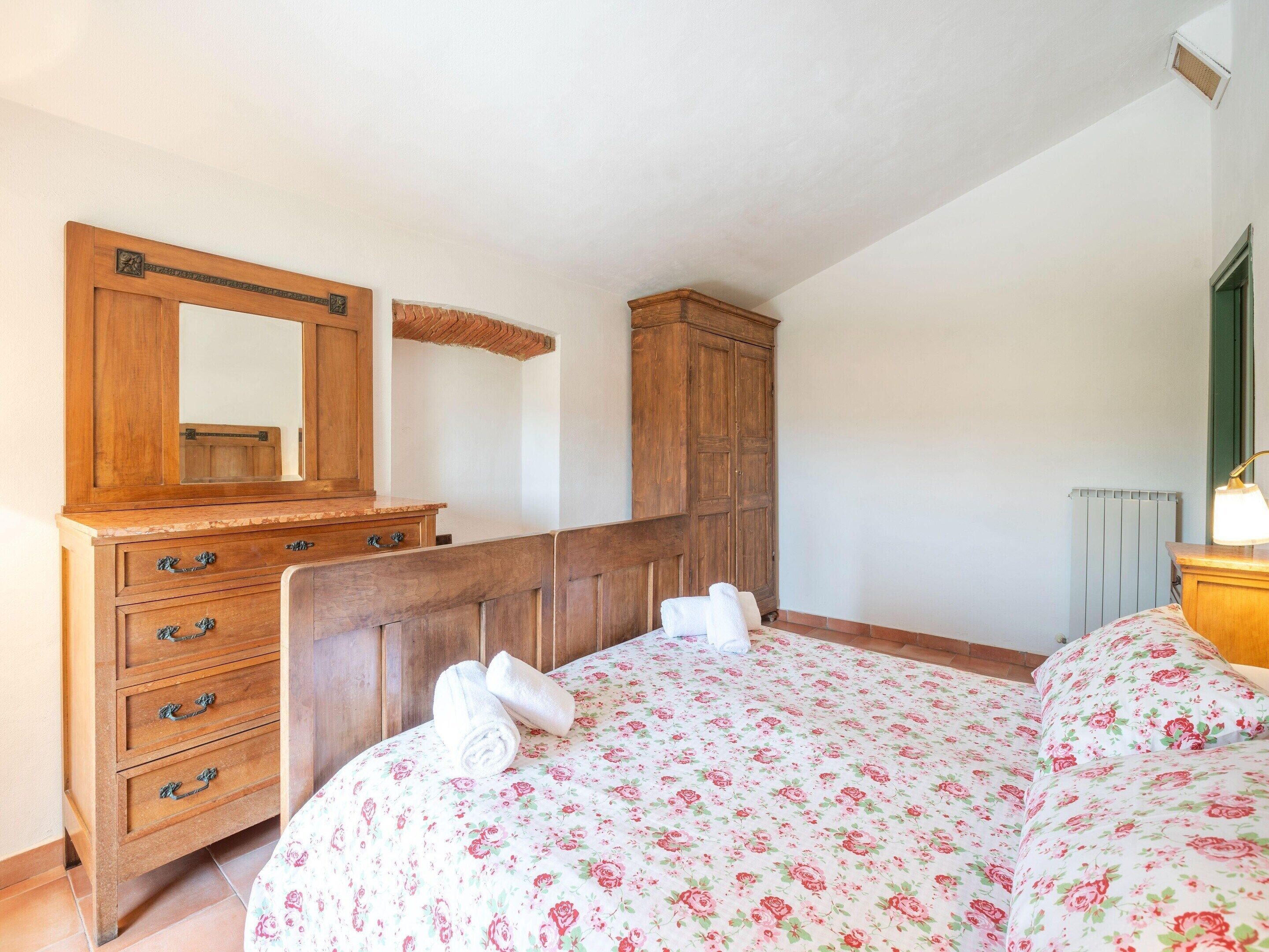 Une chambre avec une commode en bois, un grand miroir, un lit avec une literie à motifs floraux et un radiateur.