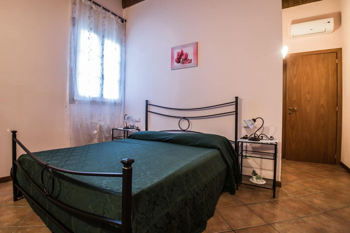 A bedroom with a bed, a nightstand, a wall-mounted air conditioner, a door, and a window with curtains.