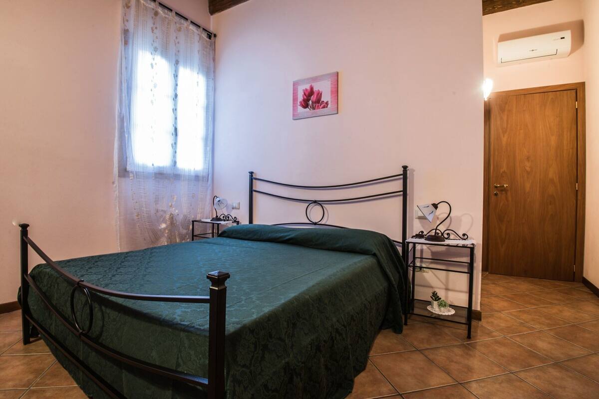 A bedroom with a bed, a nightstand, a wall-mounted air conditioner, a door, and a window with curtains.