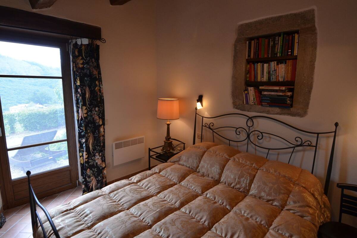 Quarto com uma cama grande, janela com cortinas, mesa de cabeceira com abajur e estante de livros.