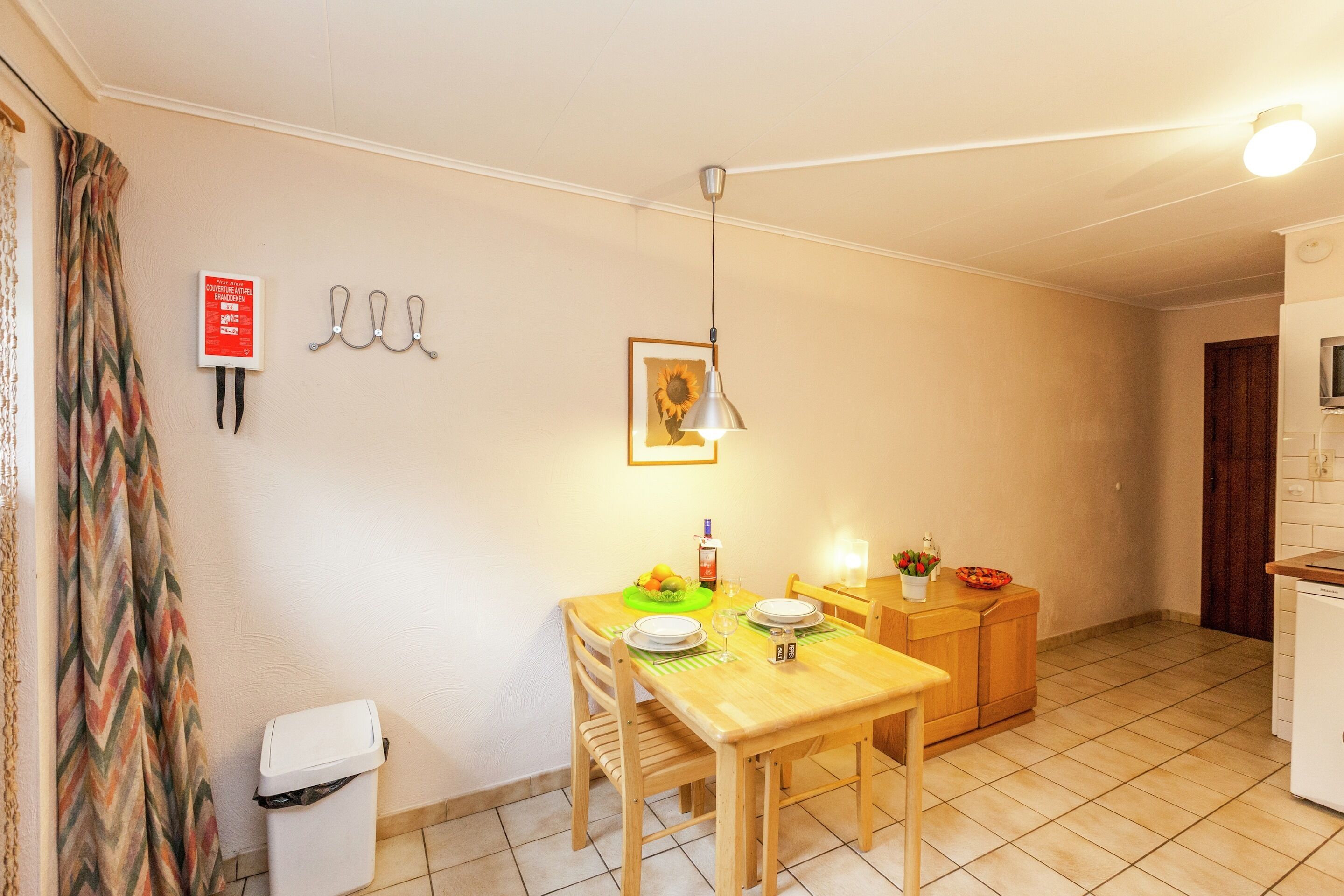 A compact kitchen with a wooden table set for dining, a trash bin, and a small cabinet.