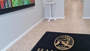 A hallway with a wooden floor, a black doormat with a logo, a small white table with a plant, and a colorful abstract painting on the wall. - HANNIBAL - Design Apartment Lilie 2 (Wolfsburg)