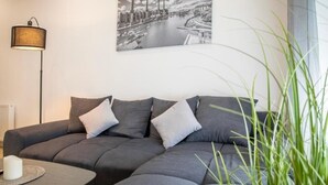 A modern living room with a grey sofa, a floor lamp, and a cityscape painting on the wall. - HANNIBAL - Design Apartment Lilie 2 (Wolfsburg)
