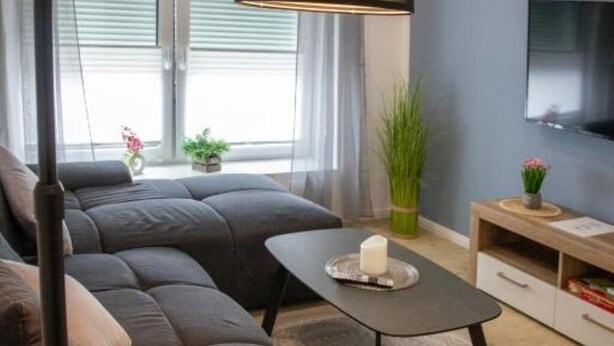A modern living room with a dark sofa, a small coffee table, and a TV mounted on the wall. - HANNIBAL - Design Apartment Lilie 2 (Wolfsburg)