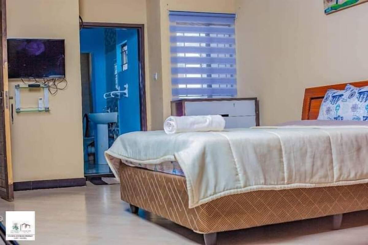 Habitación de hotel con cama, ventana con persianas, lavamanos y televisor fijado a la pared.