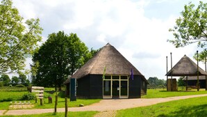 A traditional thatched-roof building with a glass entrance, surrounded by a grassy area and trees. - A Detached Bungalow With Outdoor Fireplace, Covered Terrace and Pond in a Forest Plot (Wateren)