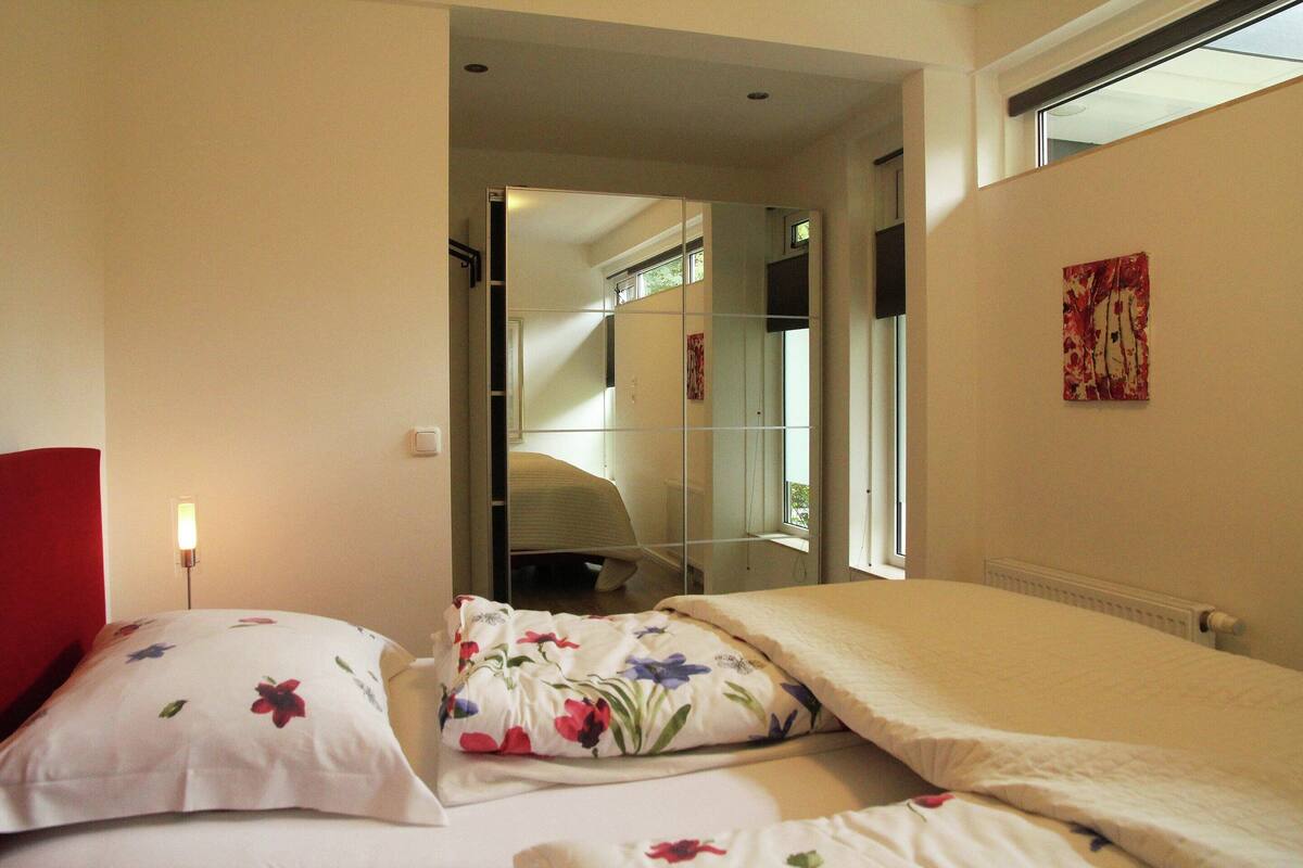 A bedroom with two beds, a red headboard, a bedside lamp, a window, and a painting on the wall.