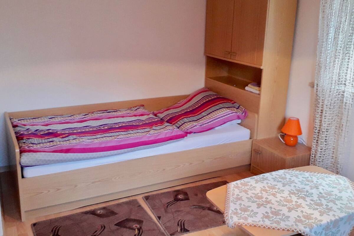A small, neatly made bed with a striped blanket, a wooden nightstand with a lamp, and a small table with a floral tablecloth.