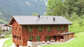 A wooden chalet with a sloped roof, large windows, and a stone staircase leading to a patio area with outdoor furniture.