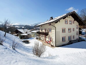Un edificio cubierto de nieve con múltiples ventanas y un balcón, rodeado de un paisaje nevado y montañas distantes.