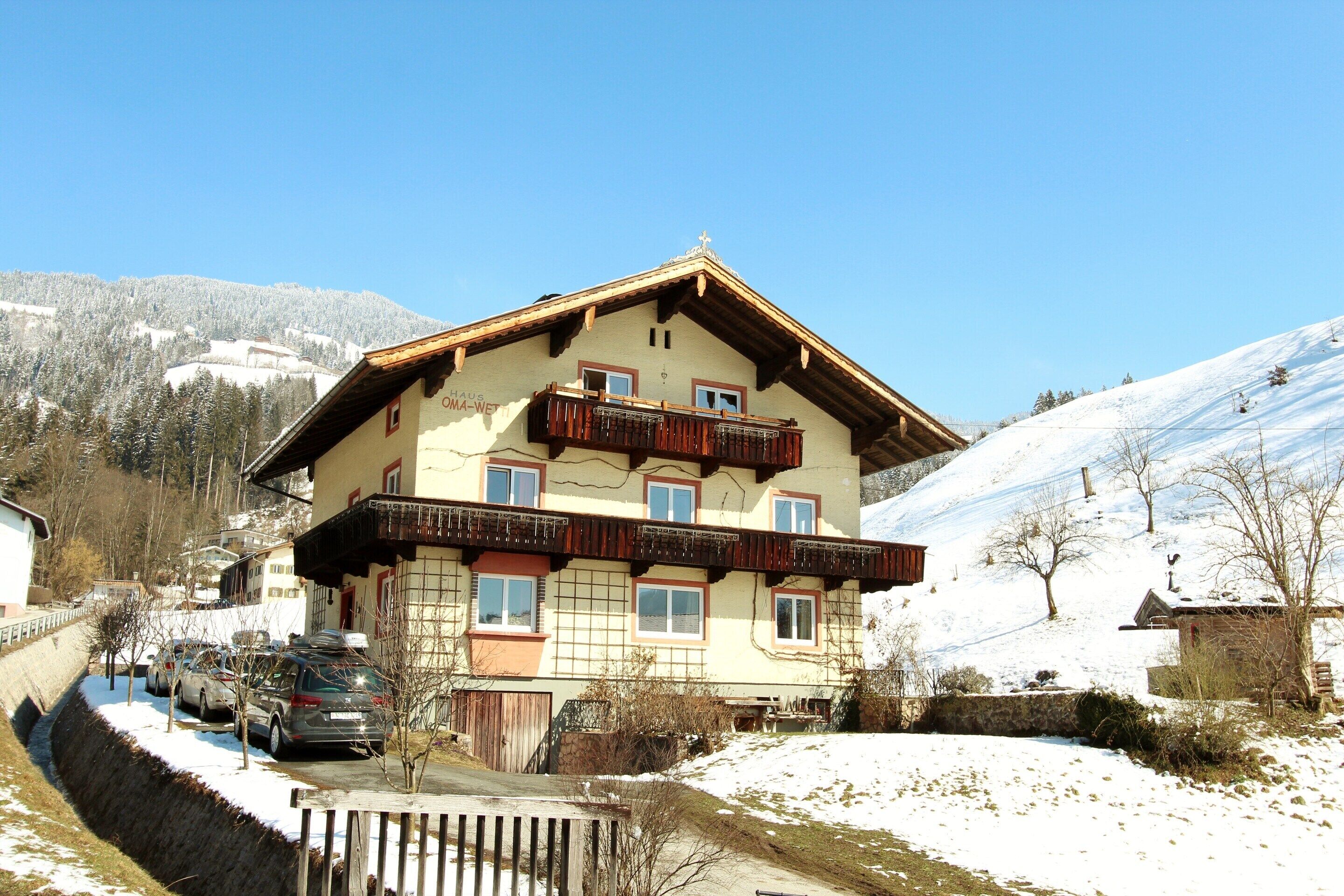 A two-story chalet with a balcony, surrounded by snow-covered hills and parked cars.