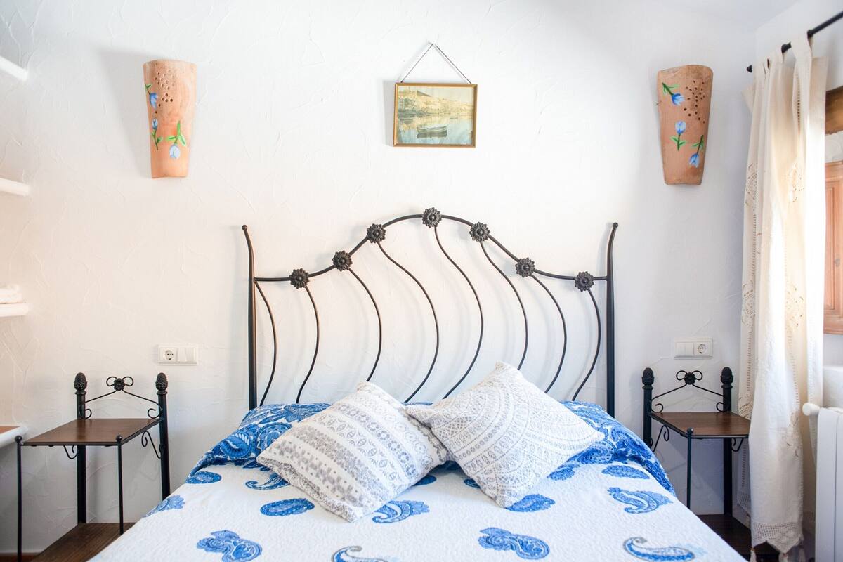 A bed with a patterned bedspread, two wall-mounted sconces, and a framed picture above the bed.
