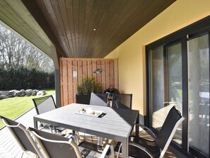 Eine Terrasse im Freien mit Tisch und Stühlen, einer Holzbalkendecke, einer Glastür und Blick auf den Garten.