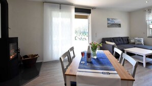 Uma sala de estar moderna com lareira, uma mesa de jantar com cadeiras, um sofá e uma grande janela com vista para o verde.