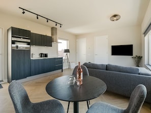 A modern living room with a dark round table, grey sofas, and a kitchen area with dark cabinets and a built-in oven.
