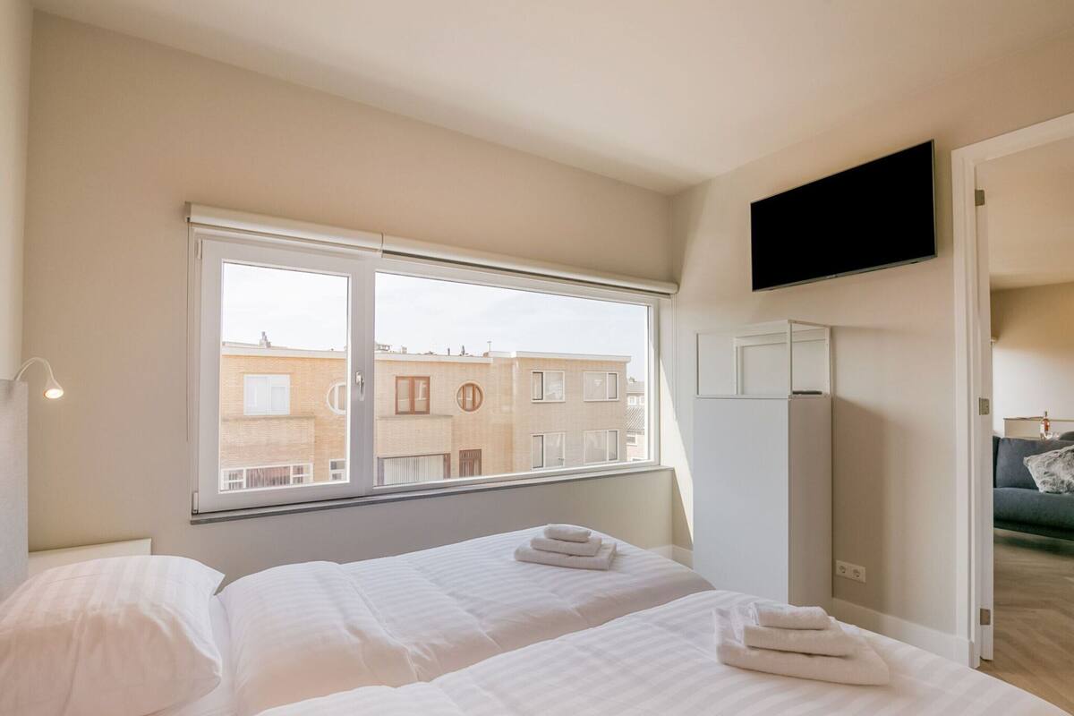 Une chambre d’hôtel avec un lit, un téléviseur fixé au mur et une fenêtre donnant sur un immeuble.