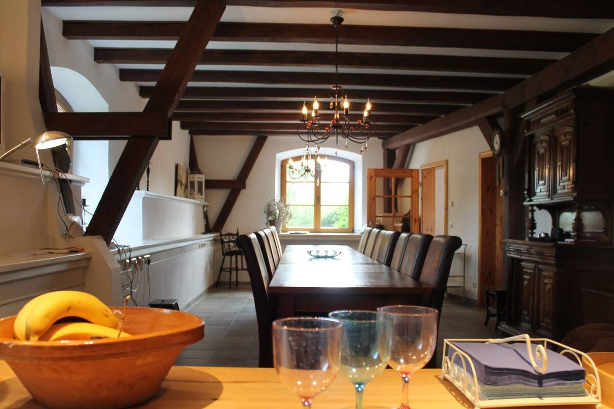 A dining area with a long table, chairs, and a chandelier. There is a bowl of fruit and wine glasses on the table.