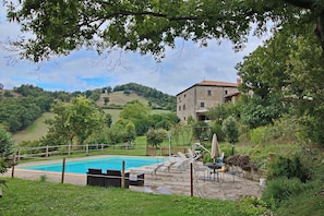 A pool area with lounge chairs and a table, surrounded by greenery and a stone building in the background. - Spacious Farmhouse in Apecchio With Pool (Apecchio)