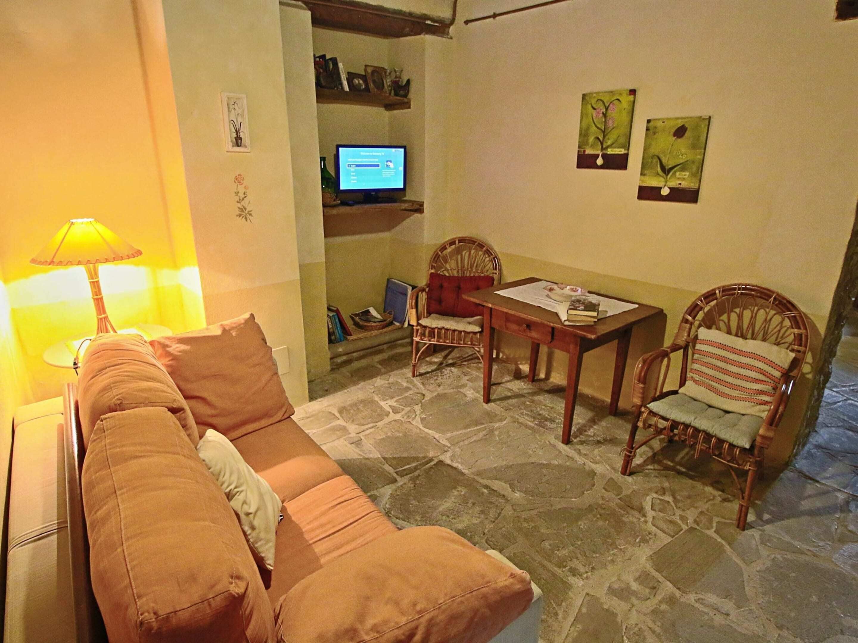 Uma sala de estar acolhedora com um sofá, uma mesa e duas cadeiras. Há uma televisão presa na parede e objetos decorativos nas prateleiras.