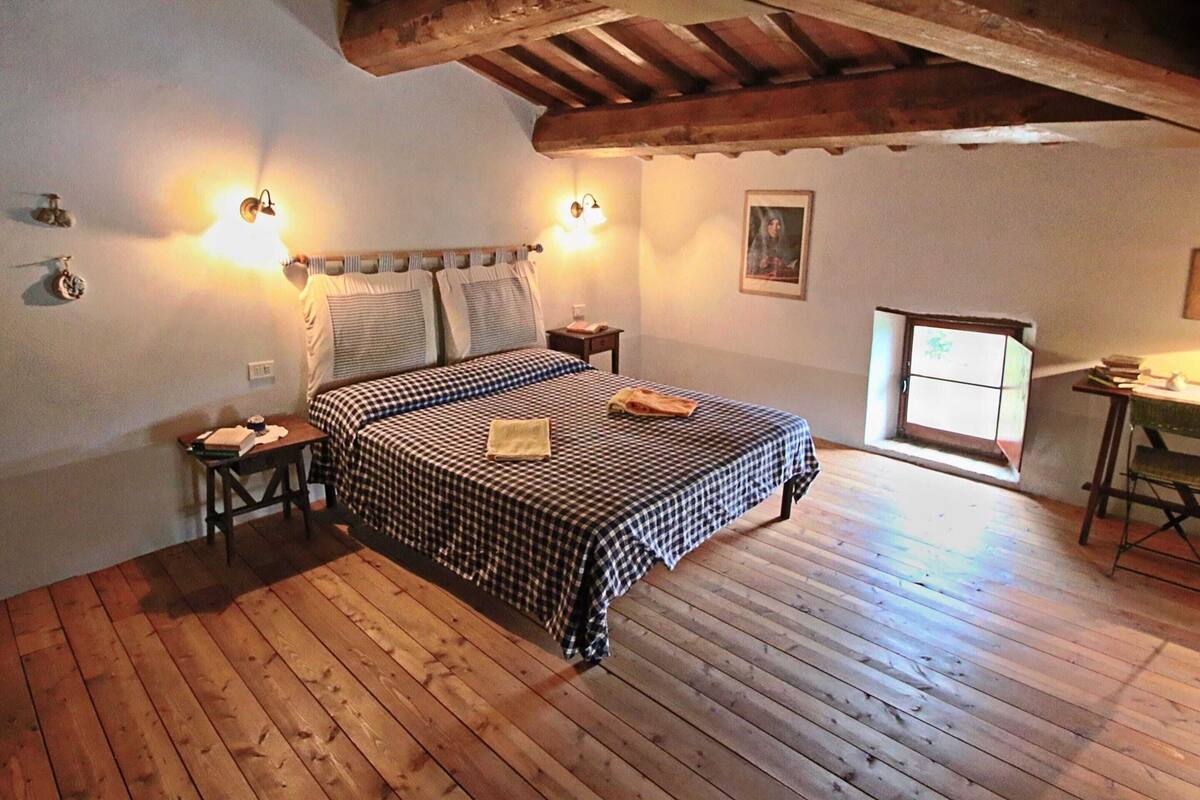 A bedroom with a wooden floor, a checkered bedspread, two bedside lamps, a nightstand, a small table with a chair, and a window with a view of the outside.