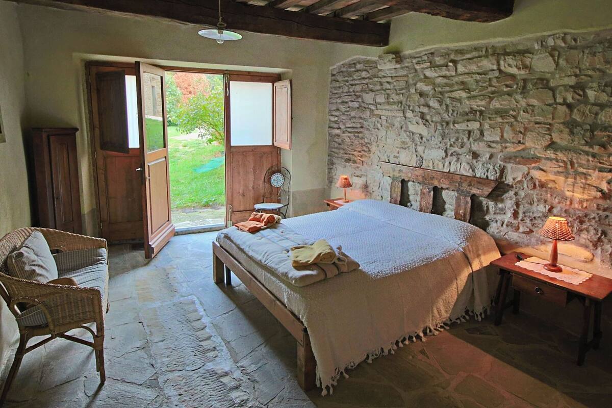 A bedroom with a stone wall, a bed, a wooden bench, a chair, and a small table with a lamp.