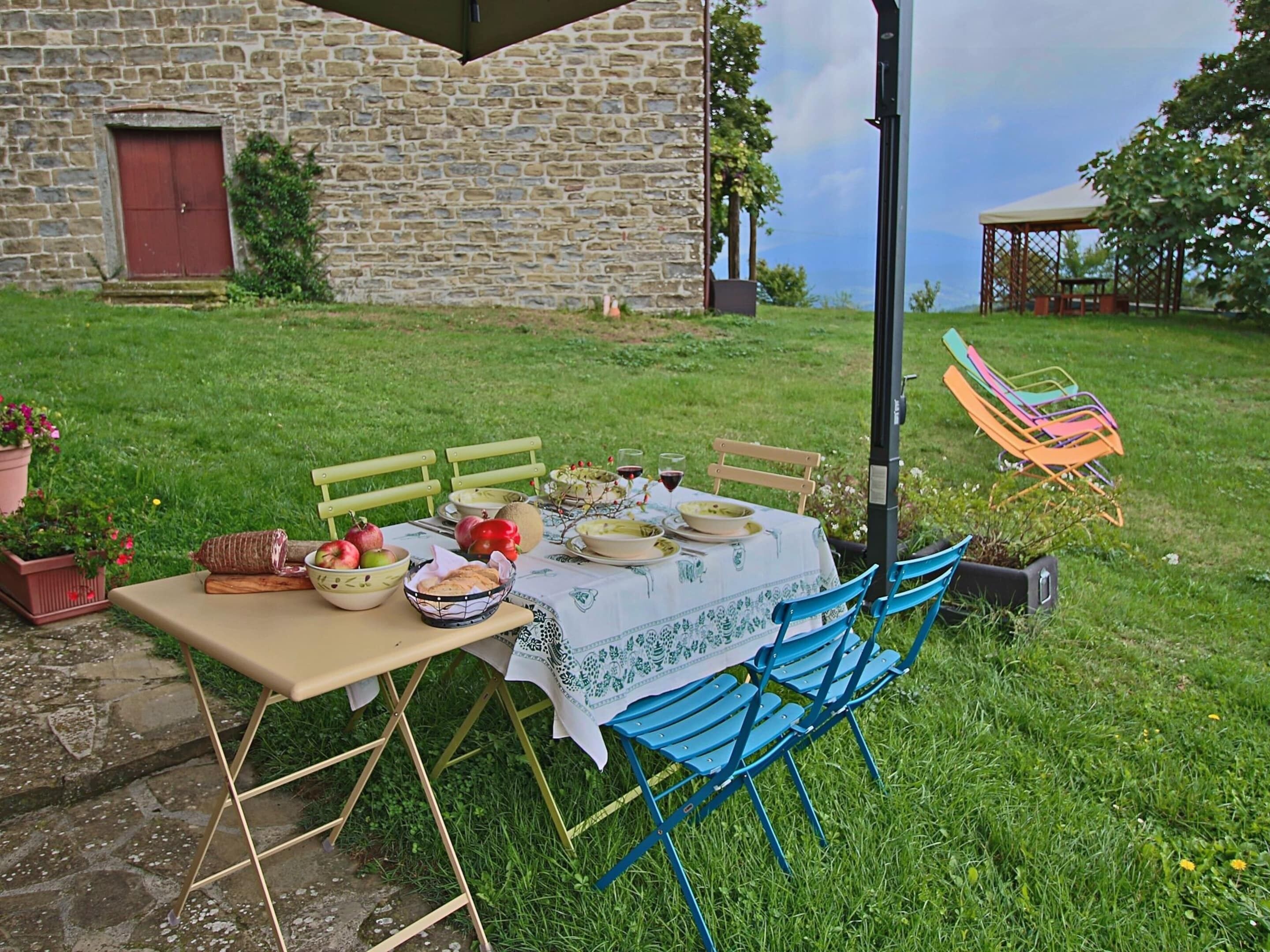 An outdoor dining setup with a table set for four, surrounded by blue chairs, in a grassy area with a stone building in the background.