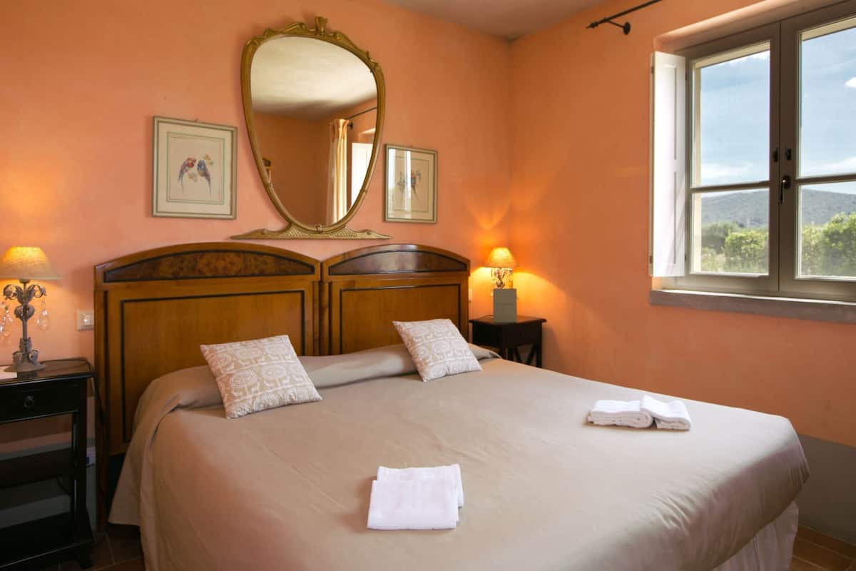 A hotel room with a large bed, two bedside lamps, a mirror, and a window with a view of greenery.