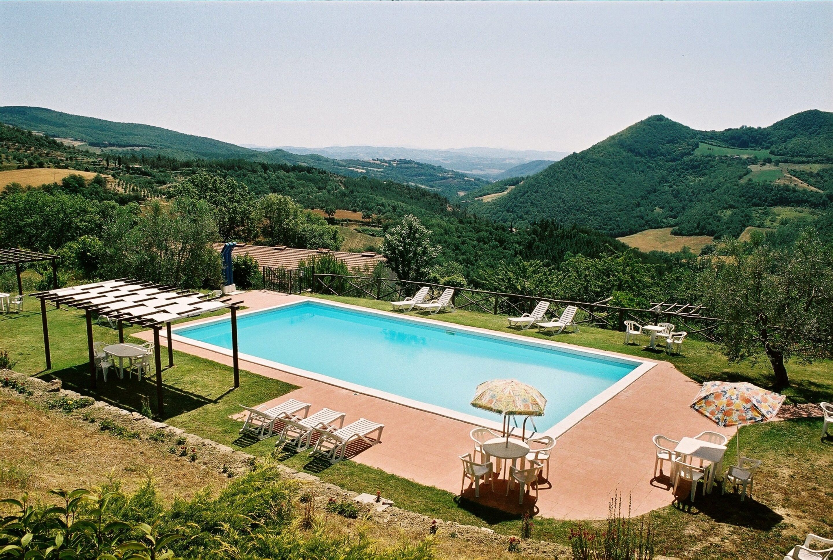 A swimming pool with lounge chairs and umbrellas, surrounded by a lush landscape and hills.