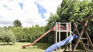 Un parque infantil con tobogán y estructura de escalada en un área verde con árboles al fondo.
