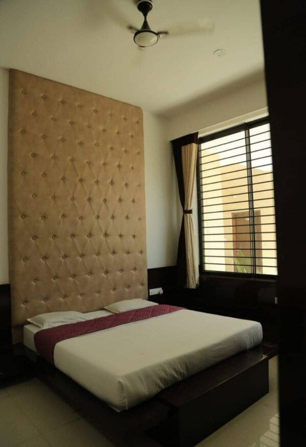 A hotel room with a large bed, a tufted headboard, a ceiling fan, and a window with curtains. - Hotel Shivani Resort (Dhar)
