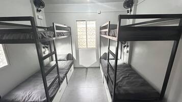 A dormitory room with bunk beds, a window with patterned blinds, and a fan on the wall.