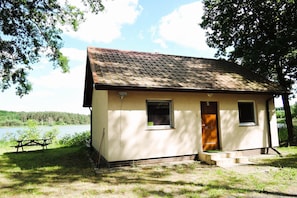 A small cabin with a tiled roof, a door, and windows, situated by a lake with a picnic table nearby. - Holiday House in Szczecin at the Lake With Parking Space for 4 Persons (Szczecin)
