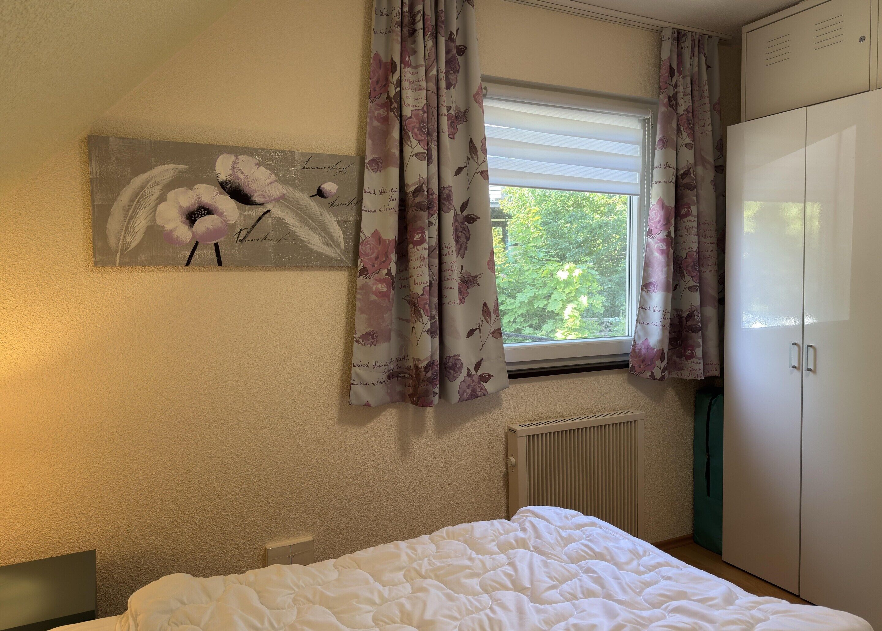 Ein Schlafzimmer mit einem Bett, einem Heizkörper, einem Fenster mit Vorhängen, einem Kleiderschrank und einem Blumenbild an der Wand.
