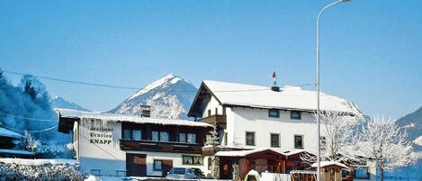 Um hotel com montanhas cobertas de neve ao fundo, uma placa visível com a inscrição 'Dachhaus Pension KNAPP' e um céu claro.