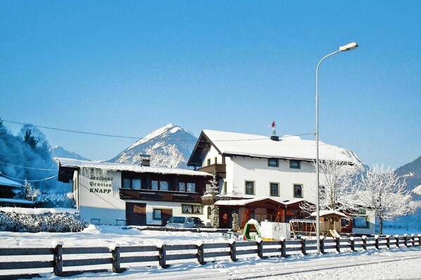 Un hôtel avec une montagne enneigée en toile de fond, une enseigne bien visible indiquant « Dachhaus Pension KNAPP », et un ciel dégagé.