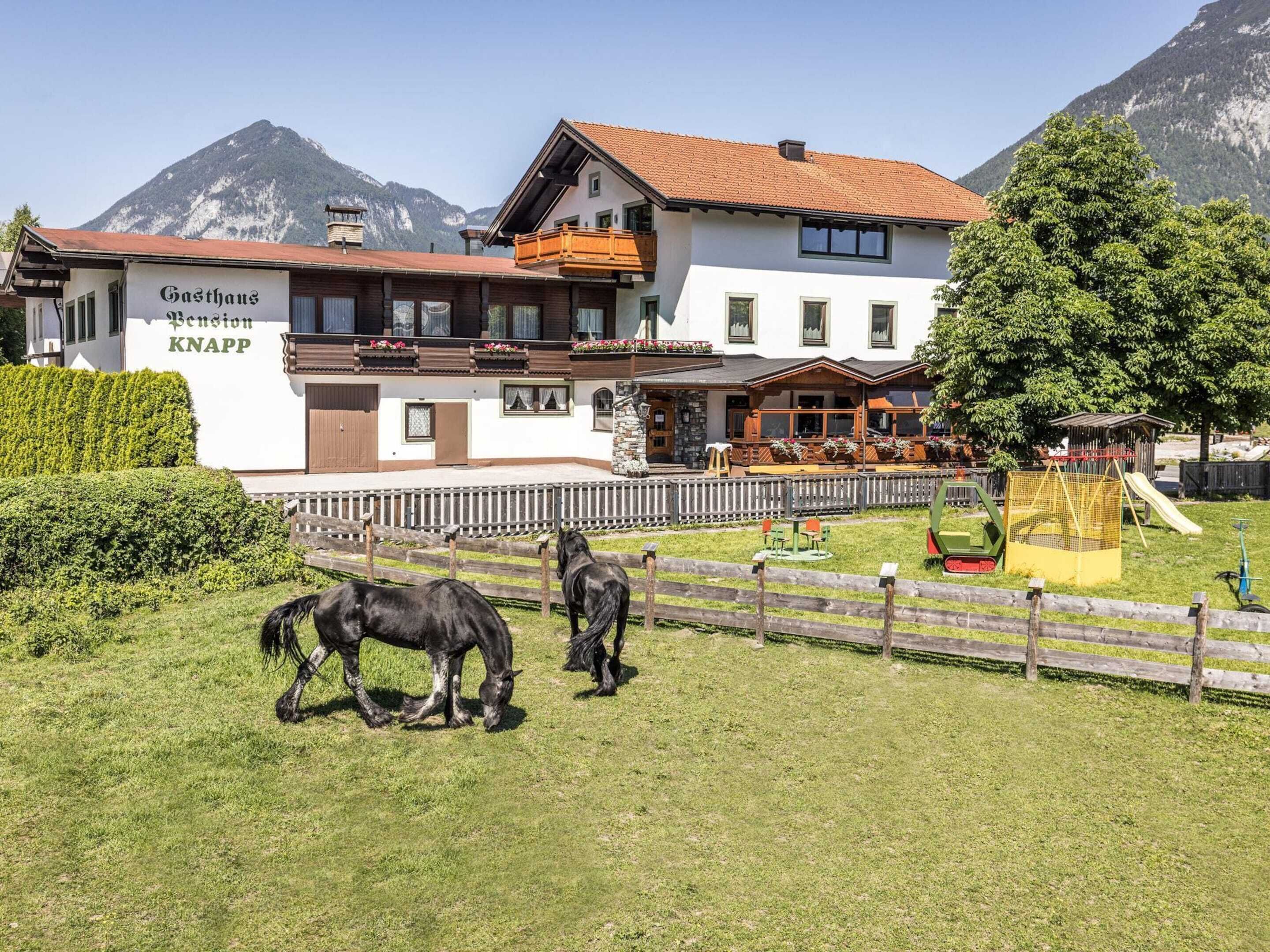 A hotel with a mountain backdrop, featuring a white facade, wooden balconies, and a fenced outdoor area with horses.