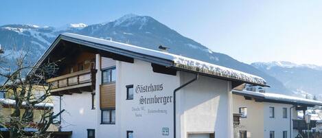 A hotel with a sign reading "Gästehaus Steinberger" and "Dusche WC" indicating a bathroom. The building has a sloped roof and is surrounded by snow-covered mountains.
