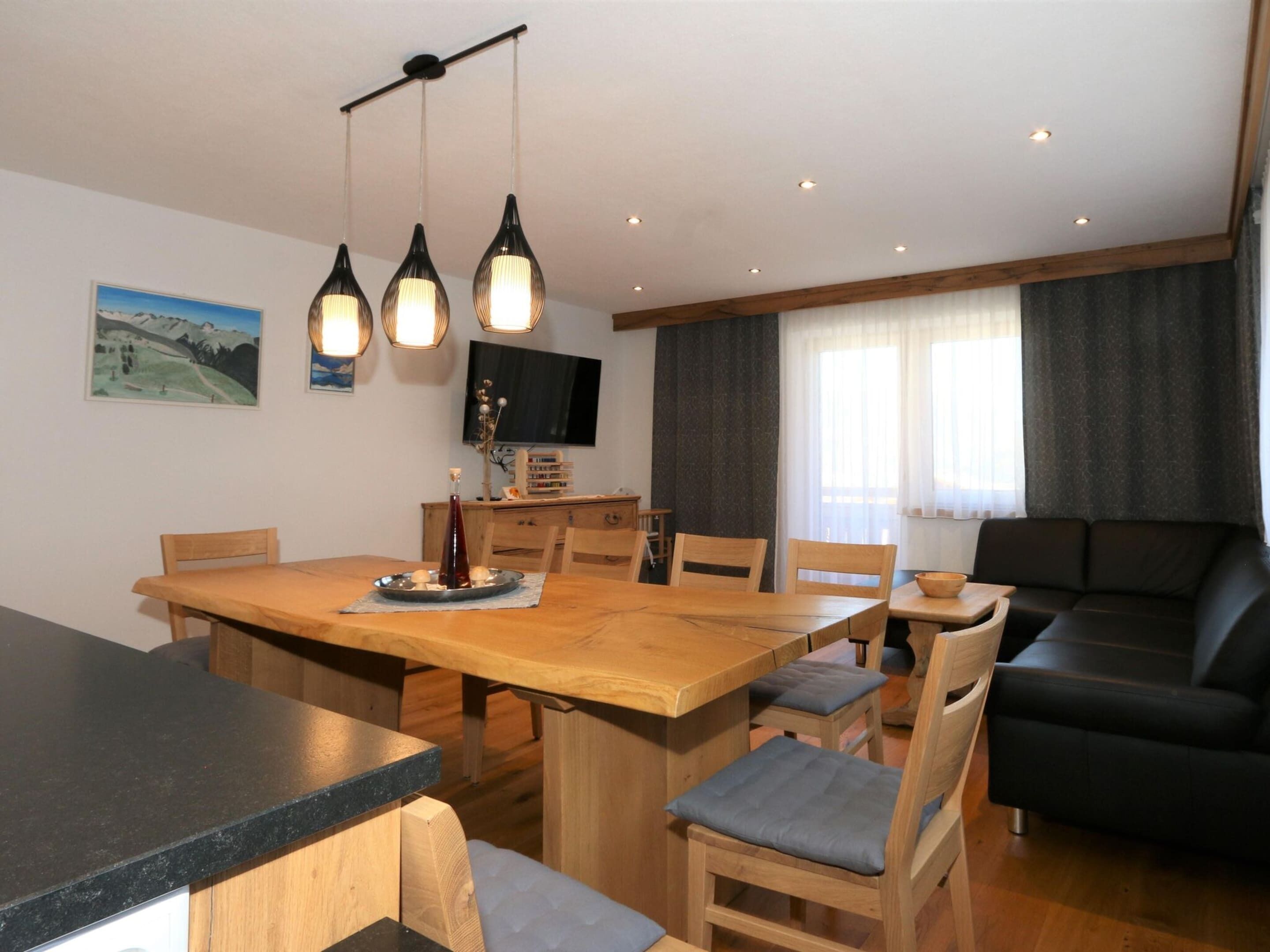 Een eethoek met een houten tafel, stoelen en hangende lampen. Achterin staat een bank en een televisie.