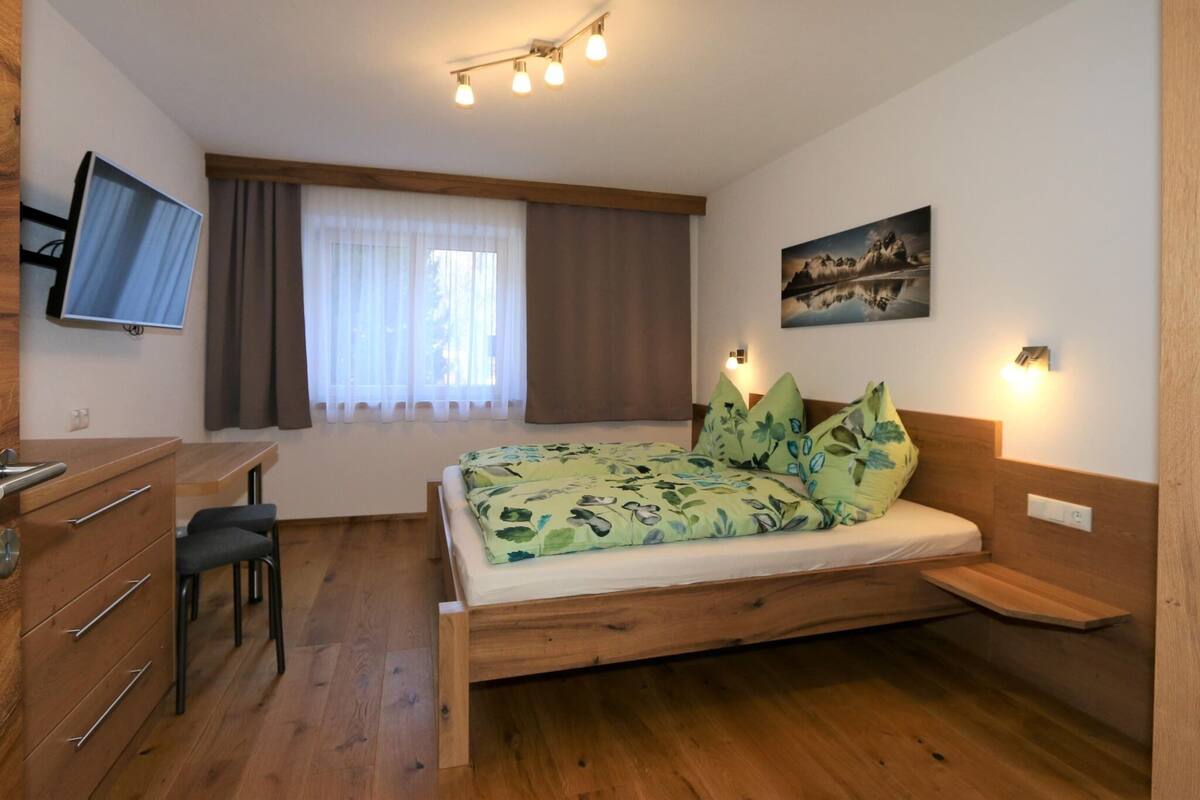A bedroom with a wooden bed, a desk with a chair, a television, and a window with curtains.