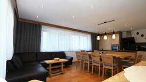 Una sala de estar moderna con un sofá negro, una mesa de comedor de madera y sillas. Se observa una cocina al fondo con estufa y armarios.