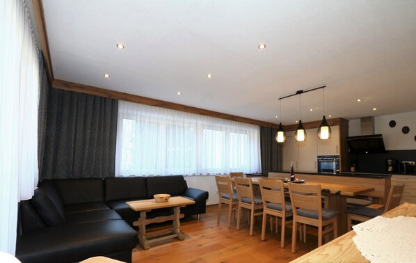 A modern living room with a black sofa, wooden dining table, and chairs. There is a kitchen area with a stove and cabinets in the background. - High-quality Holiday Apartment in Oberkrimml Near Waterfalls (Krimml)