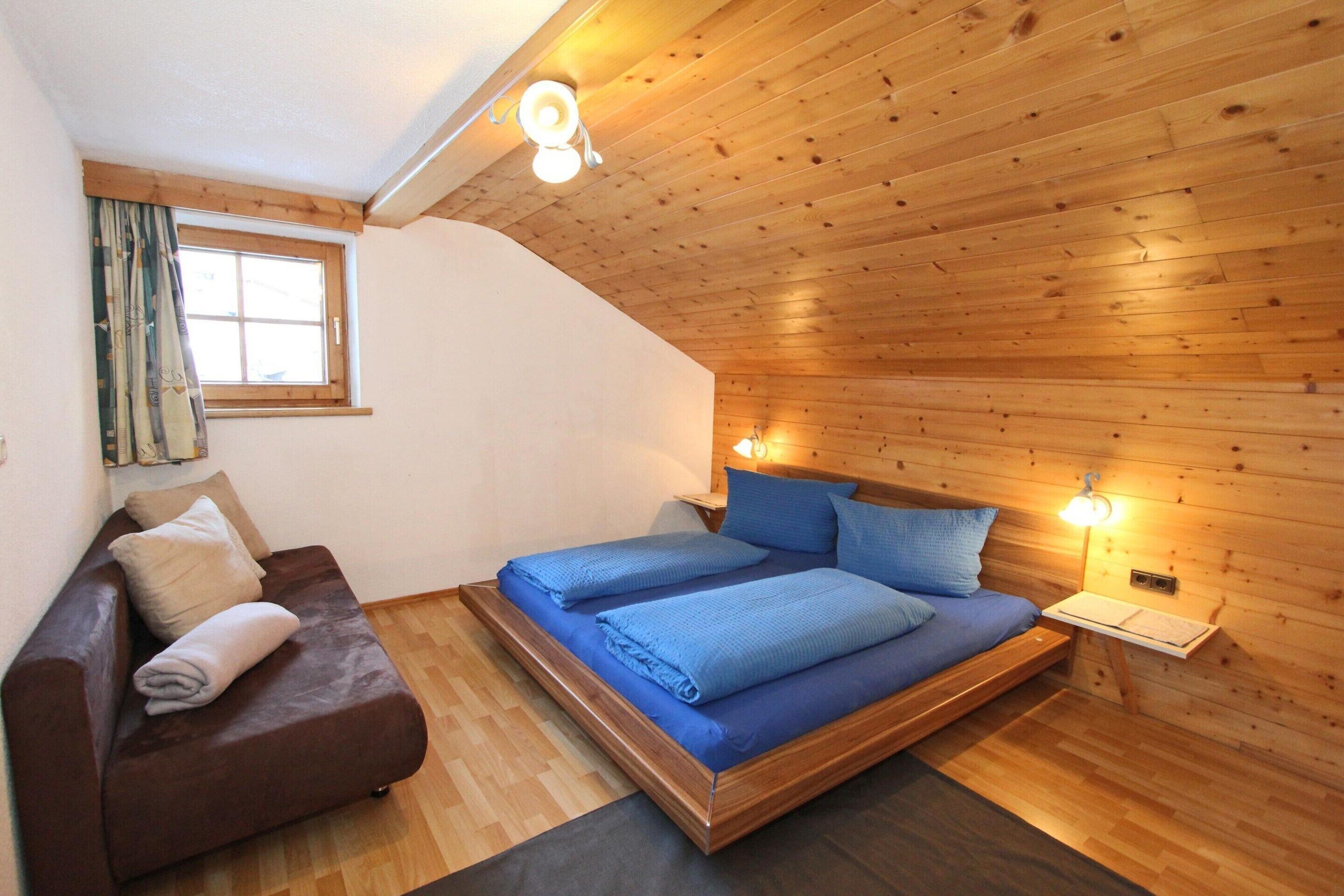 A cozy room with a bed, a wooden ceiling, a window with curtains, and a small table with a lamp.