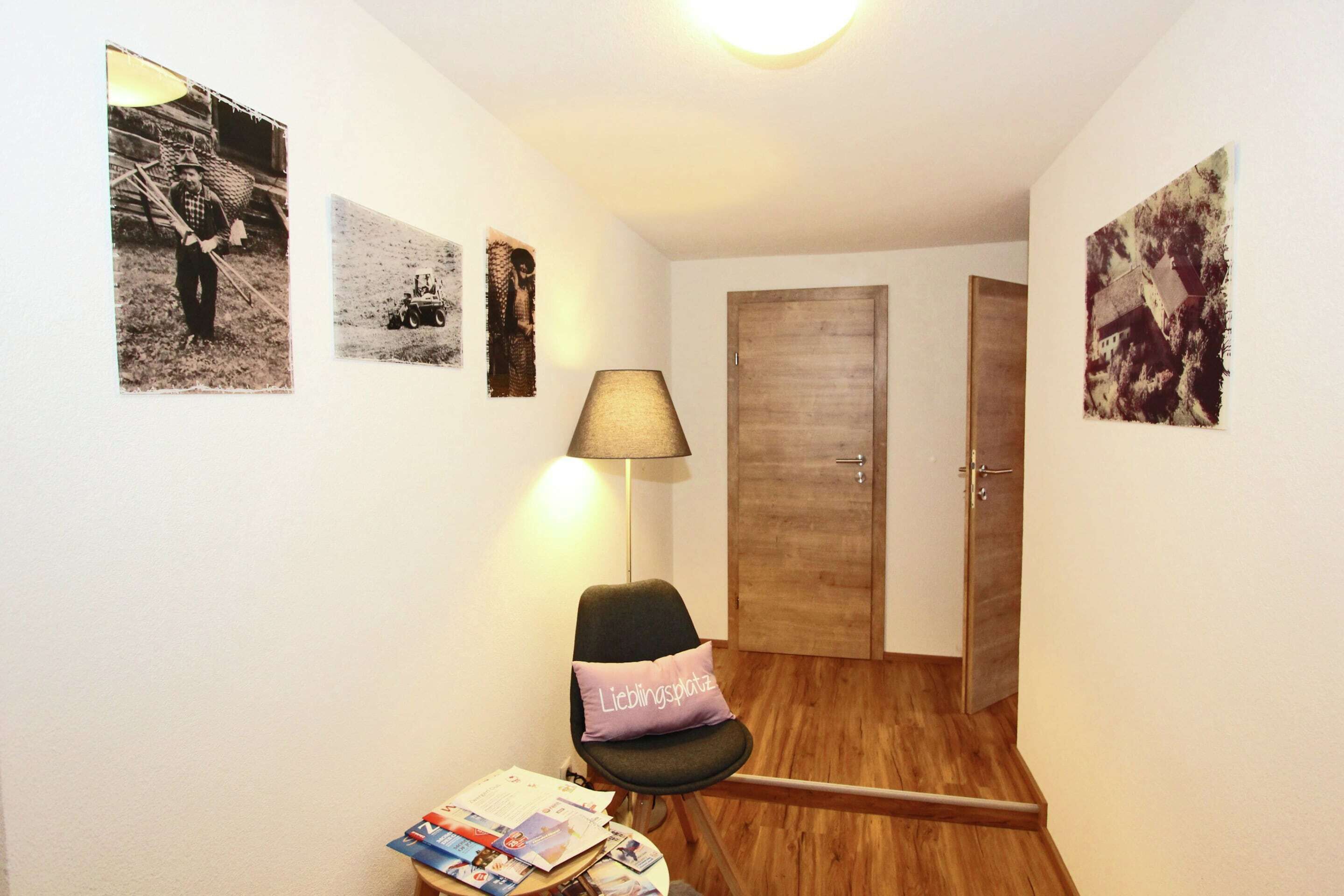 A small room with a wooden floor, a chair with a cushion, a table with magazines, and three framed pictures on the wall.
