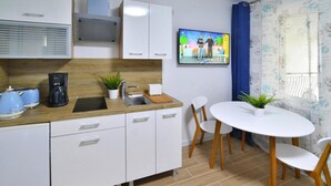Una cucina moderna con mobili bianchi, un piano di lavoro in legno, una macchina da caffè e un lavello. C'è un'area pranzo con un tavolo e sedie bianche, e una TV a parete che mostra un programma.
