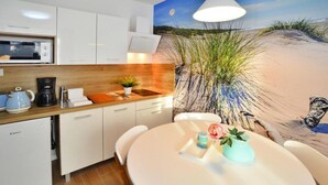 Una cucina moderna con mobili bianchi, un forno a microonde e una macchina da caffè. C'è un tavolo rotondo con tovaglia bianca, una piccola pianta e una candela. Sullo sfondo, dietro al bancone, si trova un murale con soggetto marino.